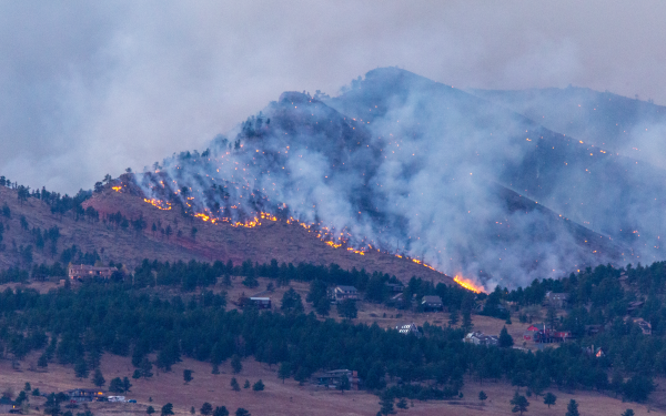 Wildfire burns over hills, moving toward homes in the valley. Smoke clouds the sky.