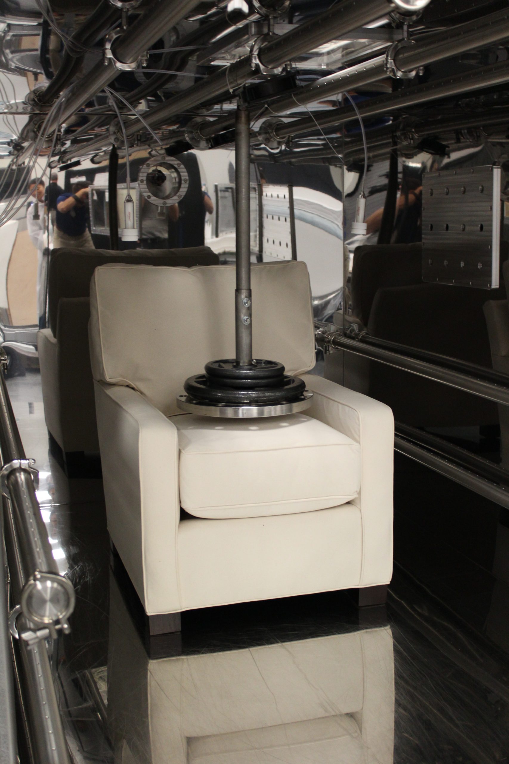 White lounge chair sits on platform in laboratory setting. Surrounded by metal piping with platform hovering over seat cushion.