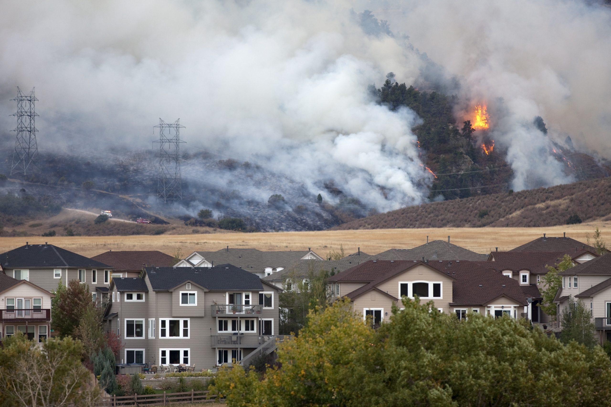 Wildfire burns in the hills and forest behind homes in foreground. Brown and yellow field sits between the hill and the homes.