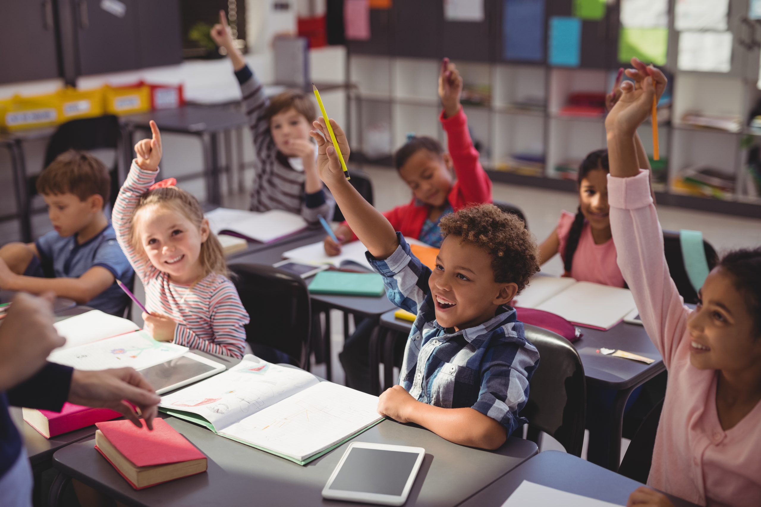 Children in classroom with their hands up, smiling at unseen person.